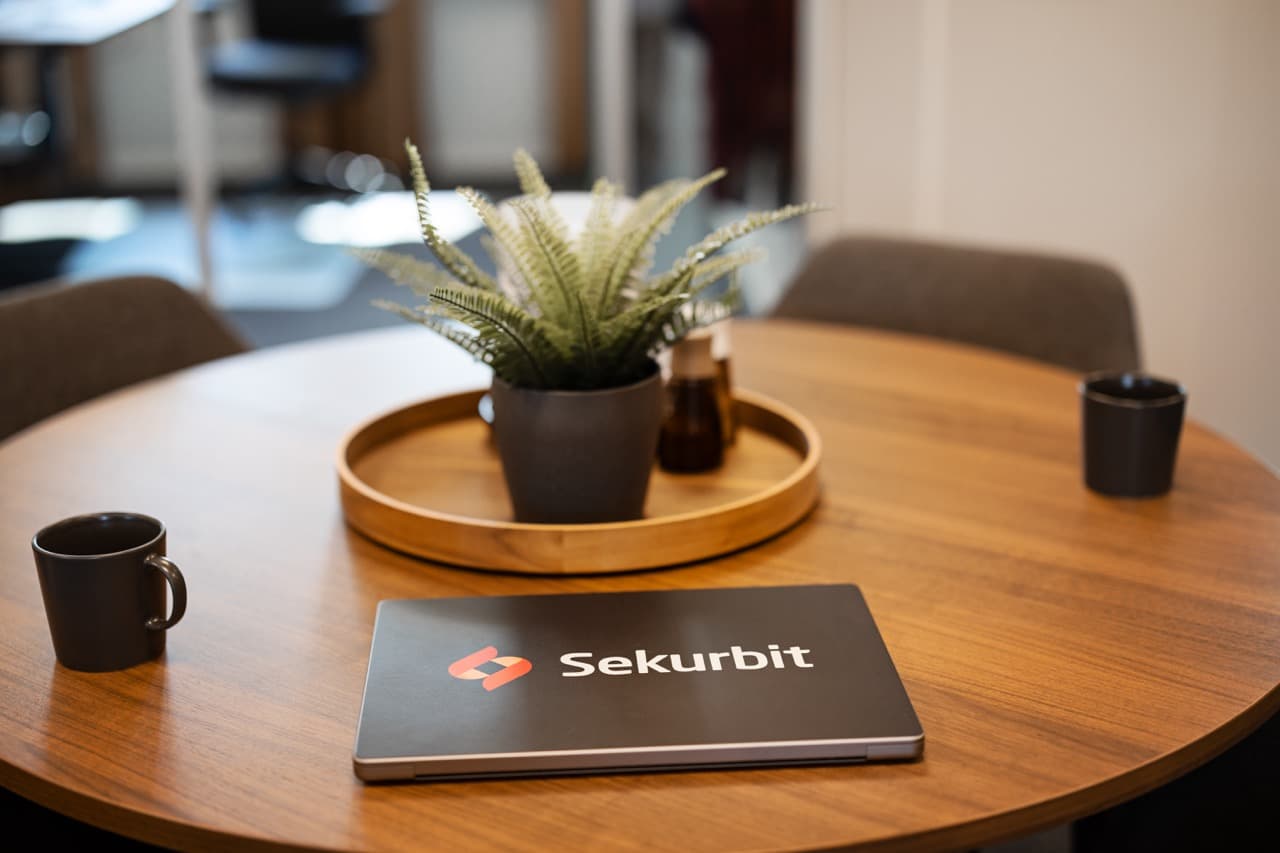 A round wooden table in an office with a potted plant on a tray, two black mugs, and a laptop displaying the Sekurbit logo.